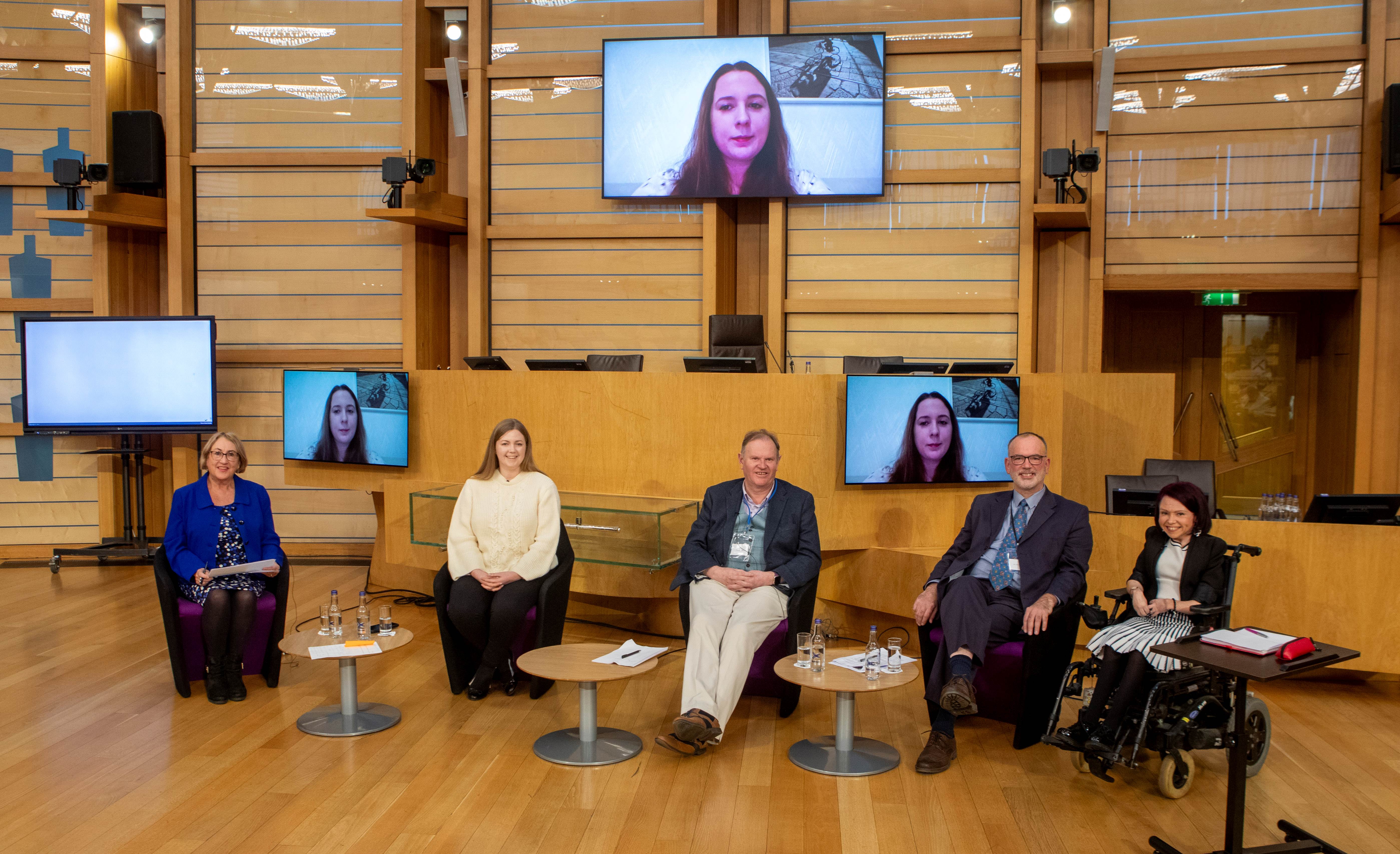 Three women and two men sit on a stage with a fourth woman pictured on screens behind them. They are smiling.